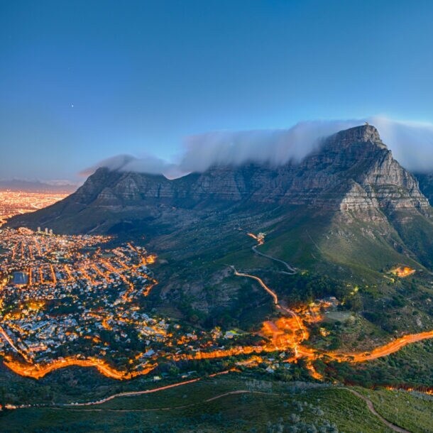 Tafelberg mit Wolkenkappe über Kapstadt bei Sonnenuntergang, beleuchtete Stadt und Küste im Hintergrund