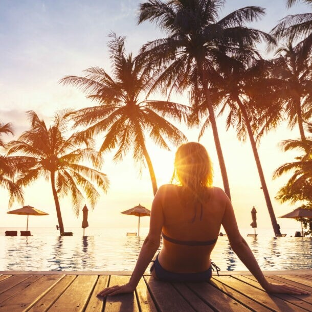 Frau sitzt am Rand eines Pools mit Blick auf Sonnenuntergang und Palmen