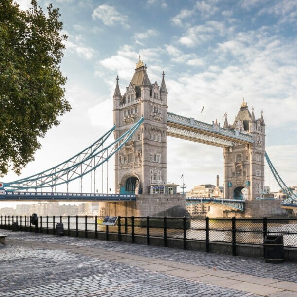 Tower Bridge über der Themse mit zwei markanten Türmen und blau-weißen Hängebrückenabschnitten, Blick vom Ufer mit Bäumen und Laterne.