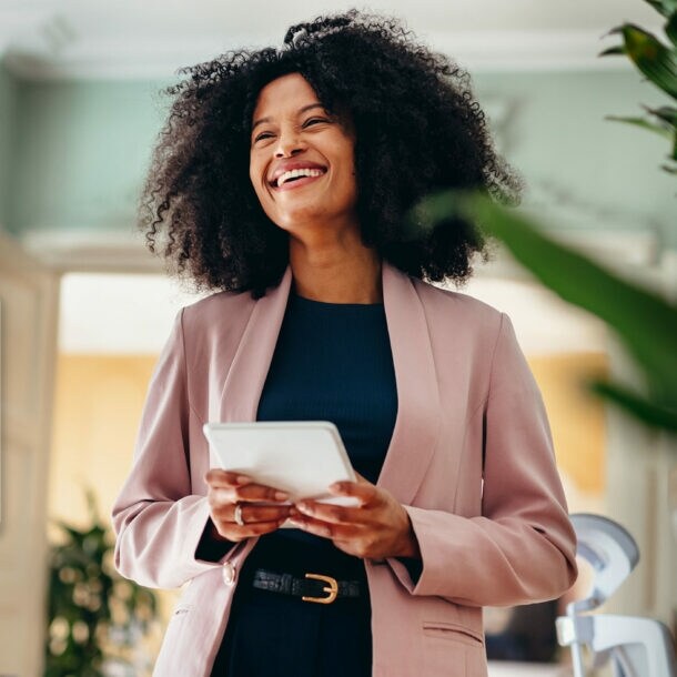 Eine lachende Frau in einem rosafarbenen Blazer steht in einem modernen Büro und hält ein weißes Tablet in den Händen, daneben ein Freisteller einer silbernen American Express Business Kreditkarte über einer einfachen American Express Platinum Card.