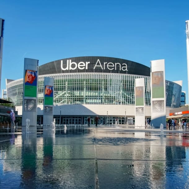 Rundes Gebäude mit Glasfassade und der Aufschrift Uber Arena hinter einem bebauten Platz mit Werbetafeln um eine Wasserfläche.