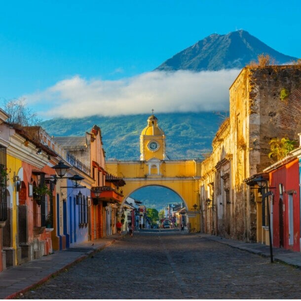 Blick auf die gelbe Santa-Catalina-Arkade in Antigua, Guatemala, mit Kopfsteinpflasterstraße und Vulkan Agua im Hintergrund