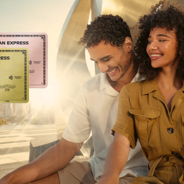 Ein lächelndes Paar sitzt vor einer gold glänzenden Metallskulptur im Sonnenschein auf einem weitläufigen Platz, er hat einen Arm um sie gelegt.