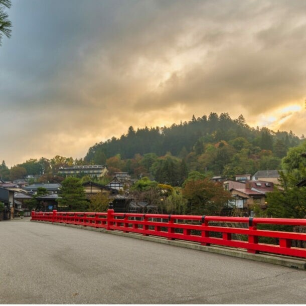 Rote Brücke über Fluss mit Häusern und bewaldetem Hügel im Hintergrund bei bewölktem Himmel