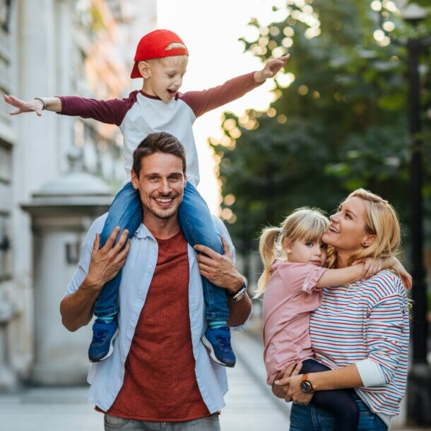Ein Mann auf einem Weg zwischen Haus und Straße trägt einen Jungen auf den Schultern, eine Frau daneben trägt ein Mädchen.