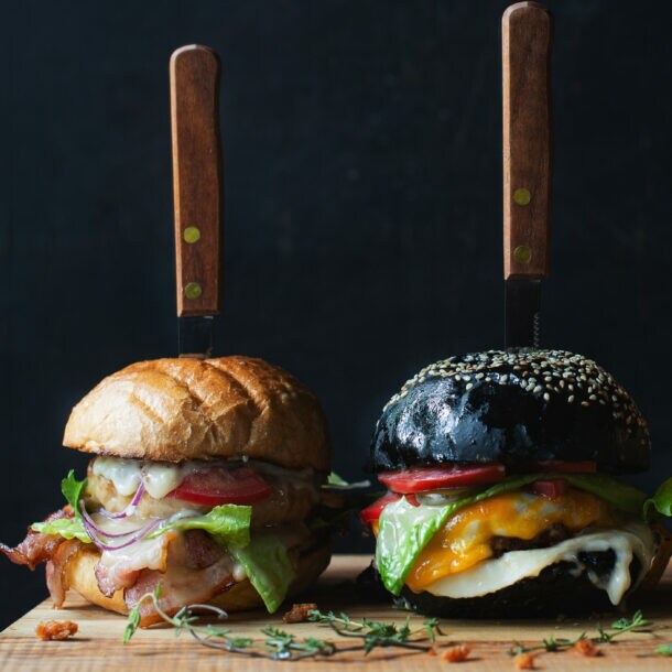 Drei verschiedene Burger mit Messern im Brötchen auf einem Holzbrett, darunter Salat, Tomate und Käse sichtbar