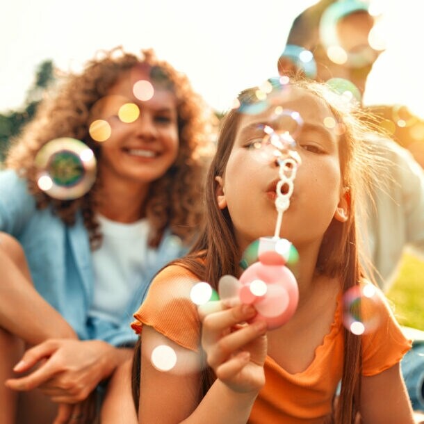 Eltern mit Kindern sitzen draußen auf Gras und spielen mit Seifenblasen bei Sonnenlicht.