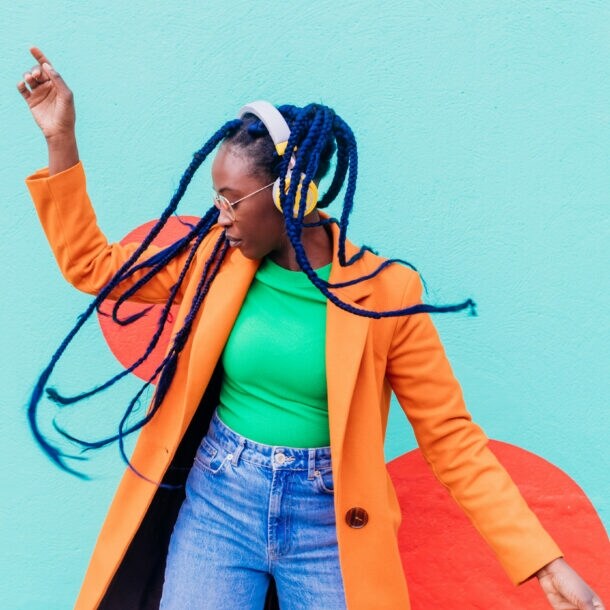 Eine junge Frau mit blauen Rastazöpfen in einem grünen Shirt und orangefarbenen Mantel tanzt mit gelben Kopfhörern vor einer türkisfarbenen Fassade.