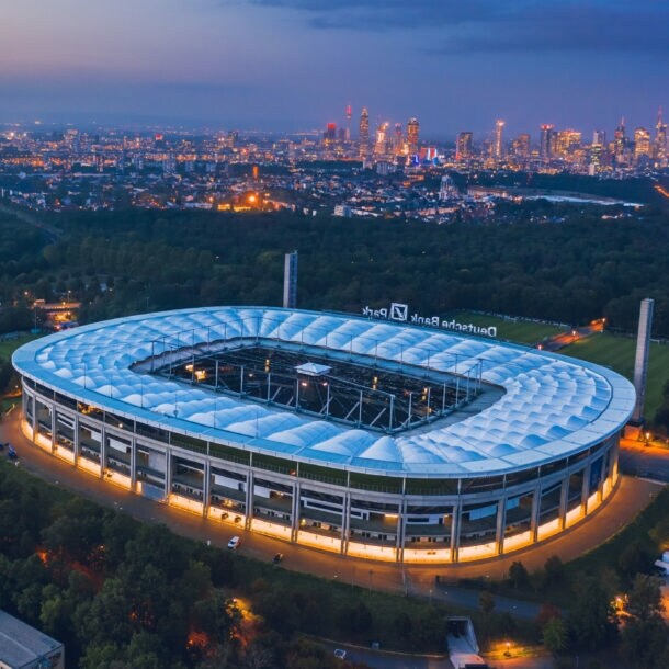 Luftaufnahme eines Fußballstadions, umgeben von Waldgebiet, vor der erleuchteten Skyline Frankfurts bei Nacht.