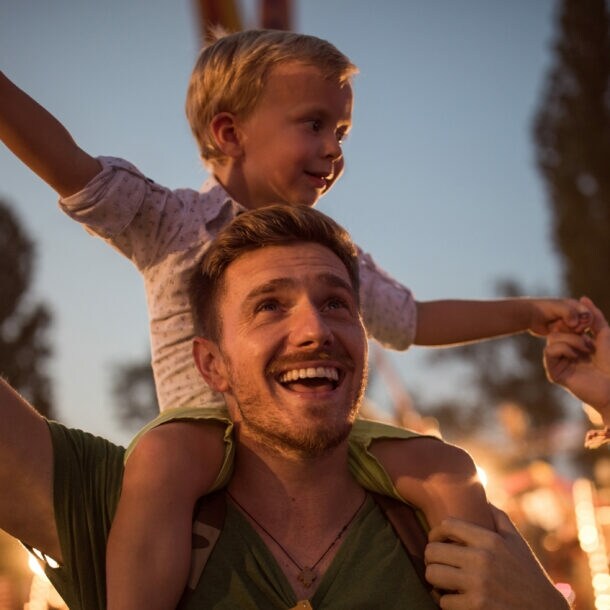 Mann im Dunkeln mit kleinem Kind auf den Schultern, im Hintergrund sind Bäume und Beleuchtung zu sehen.