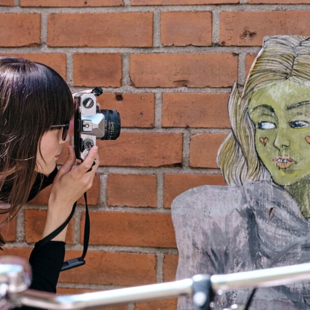 Frau mit braunen Haaren und rotem Blumenmuster-Oberteil fotografiert eine Wandzeichnung einer Person mit langen Haaren vor einer Backsteinmauer.