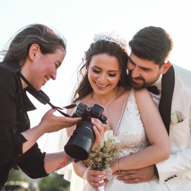 Braut in weißem Spitzenkleid mit Blumenstrauß, Bräutigam in weißem Anzug umarmt sie von hinten, Fotografin zeigt Kamera.