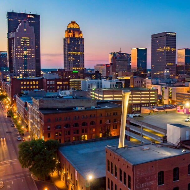 Skyline von Louisville, Kentucky, bei Sonnenuntergang mit leuchtenden Hochhäusern, historischen Backsteingebäuden im Vordergrund und der beleuchteten Innenstadtstraße.