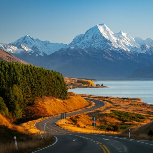 Malerischer Blick auf schneebedeckte Berge vor klarem blauem Himmel, im Vordergrund verläuft eine verlassene Straße zwischen grünen Nadelwäldern und einem ruhigen See.