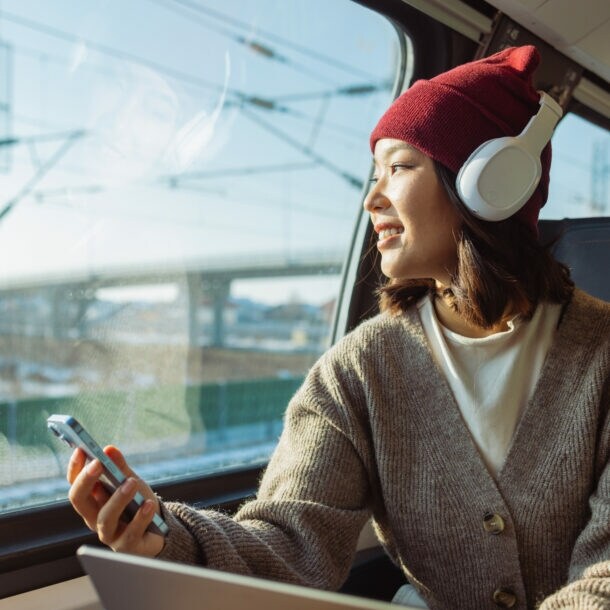 Eine Person mit Kopfhörer und Smartphone sitz in einem Zug. Sie trägt eine rote Mütze und einen grauen Pullover. Im Hintergrund sind Züge und Schienen zu sehen.