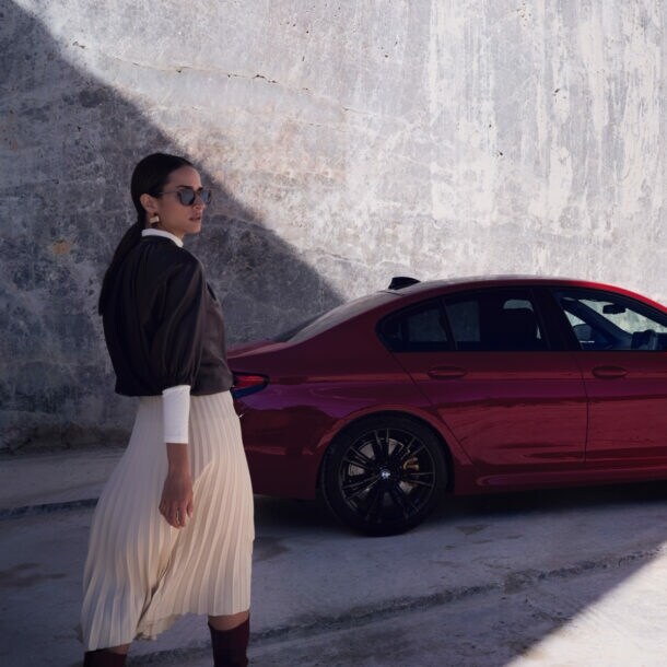 Eine elegante Frau mit Sonnenbrille vor einem roten BMW vor einer Betonwand.