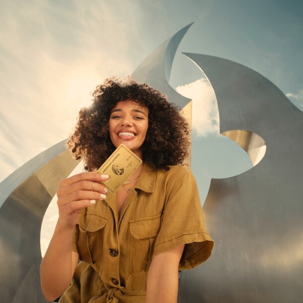 Eine lächelnde junge Frau hält eine goldene Kreditkarte von American Express in der Hand, im Hintergrund eine moderne Skulptur im Freien bei Sonnenschein.