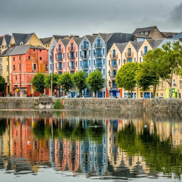 Bunte Wohngebäude in verschiedenen Farben spiegeln sich im ruhigen Wasser eines Hafens. Bäume stehen entlang der Uferpromenade.
