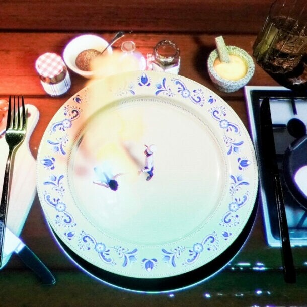 Ein gedeckter Tisch mit einem großen weißen Teller, auf dem zwei kleine Figuren stehen. Daneben befinden sich ein Messer, eine Gabel, ein Glas, Gewürzgläser und eine Pfanne mit einem Ei.