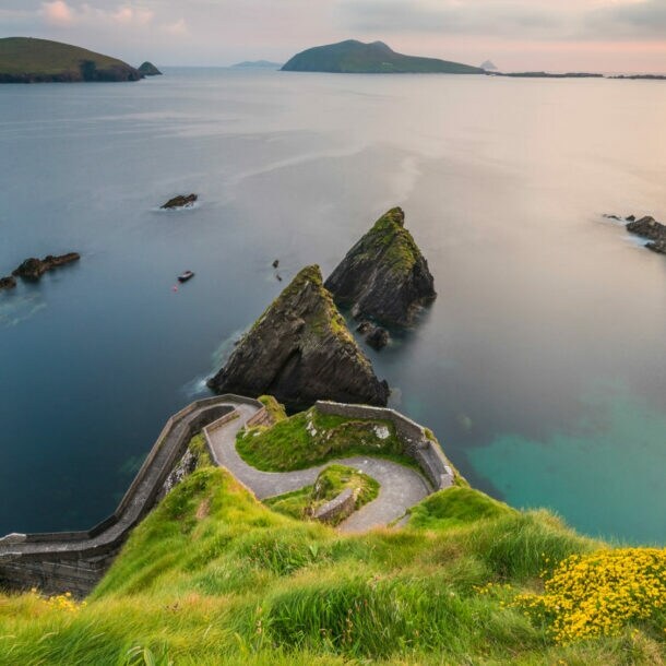 Blick auf eine Küstenlandschaft mit grünem Hügel, steilen Klippen und ruhigem Wasser. Im Hintergrund sind mehrere Inseln und ein Boot sichtbar.