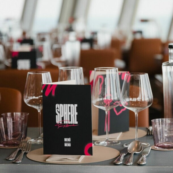 Tischgedeck in einem Restaurant mit Gläsern, einer Flasche und einer Speisekarte mit dem Titel 'SPHERE'. Im Hintergrund sind weitere Tische sichtbar.