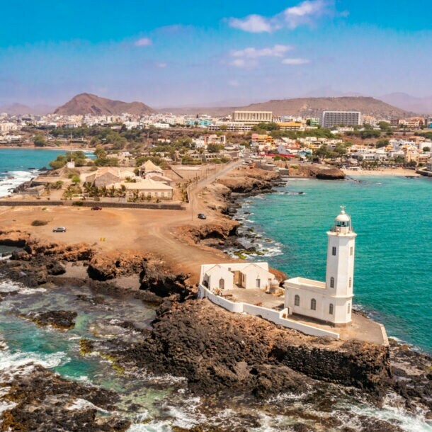 Luftaufnahme eines Leuchtturms auf einer felsigen Küste, umgeben von türkisblauem Wasser und Wellen, mit einer Stadt im Hintergrund.
