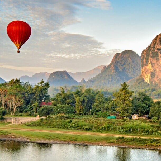 Ein roter Heißluftballon schwebt über einer grünen Landschaft mit Bergen und einem ruhigen Fluss.