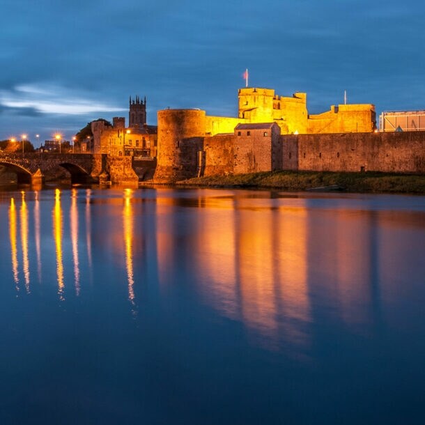 Blick auf eine beleuchtete Burg am Wasser bei Dämmerung. Die Burg hat Türme und eine Brücke im Vordergrund. Reflektionen im Wasser sind sichtbar.