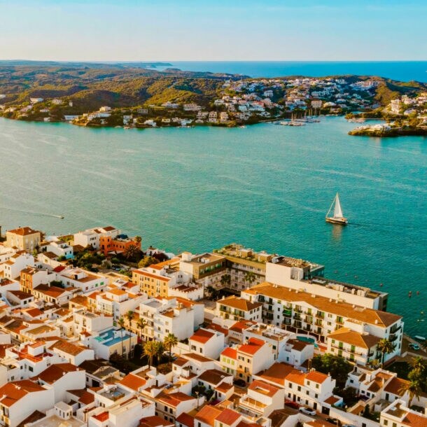 Luftaufnahme einer Küstenstadt mit weißen Gebäuden und roten Dächern, umgeben von Wasser und Hügeln. Ein Segelboot ist auf dem Wasser sichtbar.