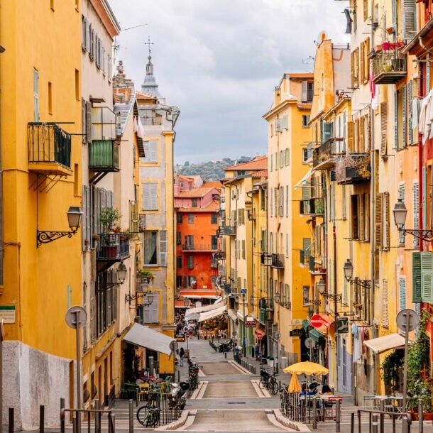 Blick auf eine enge, gepflasterte Straße mit bunten Gebäuden in Gelb- und Orangetönen. Auf beiden Seiten stehen Balkone mit Pflanzen. Im Hintergrund sind Tische und Stühle eines Cafés zu sehen.