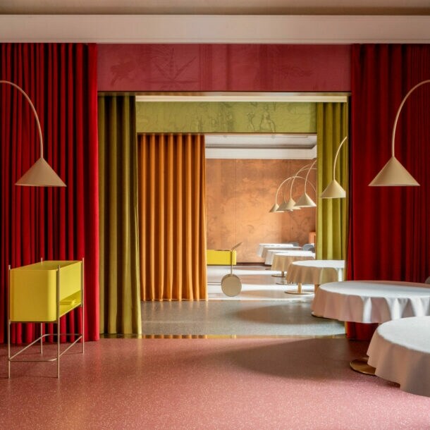 Ein eleganter Innenraum mit runden Tischen, die mit weißen Tischdecken bedeckt sind. An den Wänden hängen rote und grüne Vorhänge. Im Hintergrund sind gelbe und orangefarbene Wände sichtbar.