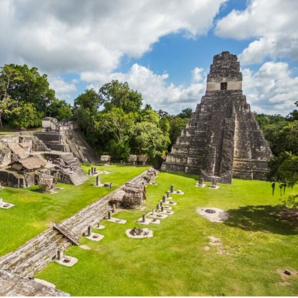 Maya-Tempelanlage mit Stufenpyramide und mehreren Steinstrukturen umgeben von grünem Gras und Bäumen unter blauem Himmel mit Wolken