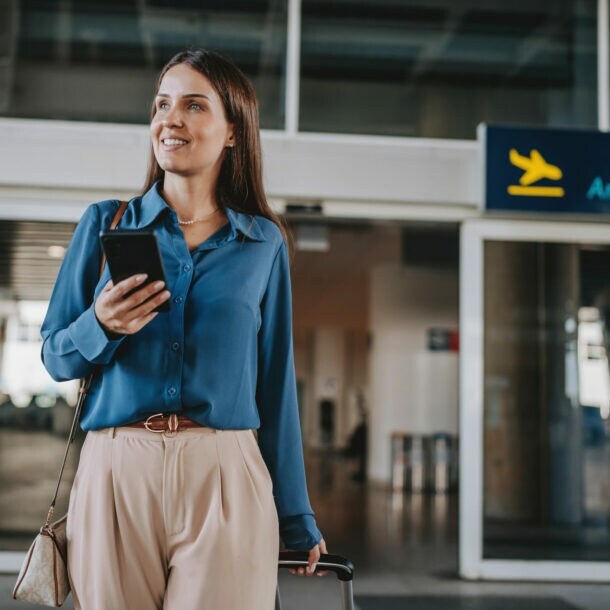 Eine elegant gekleidete Frau mit Smartphone steht vor dem Eingang der Ankunftshalle eines Flughafen-Terminals.