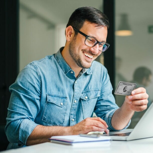 Ein gut gelaunter Mann im Jeanshemd sitzt an einem Tisch vor einem Laptop in einem Büro und hält die Business Platinum Card von American Express in der Hand.