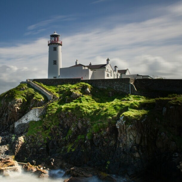 Ein Leuchtturm mit rotem Licht auf einem Hügel, umgeben von grünem Gras und steiniger Küste, mit Wellen, die gegen die Felsen schlagen.