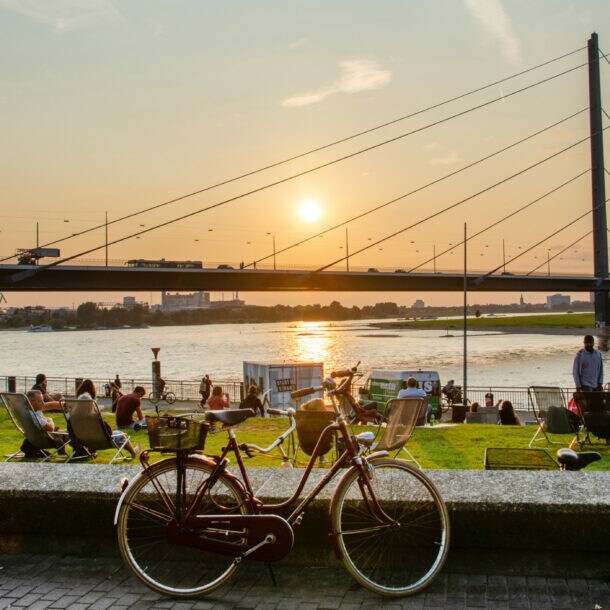Ein rotes Fahrrad steht auf dem Bürgersteig, im Hintergrund ist eine Brücke und der Sonnenuntergang über dem Wasser zu sehen. Menschen entspannen auf der Wiese.