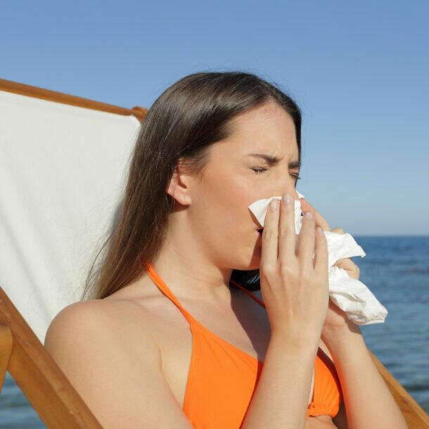 Eine Frau in einem orangefarbenen Bikini sitzt auf einer Liege am Strand und hält ein Papiertaschentuch in der Hand. Im Hintergrund sind Felsen und das Meer sichtbar.
