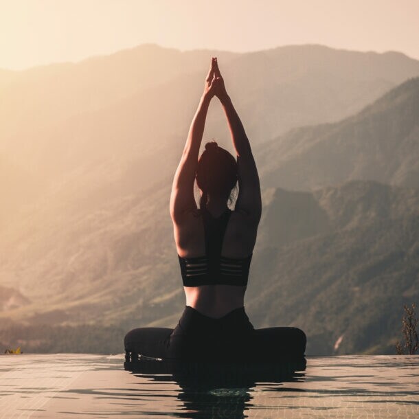 Eine Person in schwarzer Sportbekleidung sitzt im Lotussitz am Rand eines Infinity-Pools. Sie hat die Hände über dem Kopf zusammengelegt und blickt auf die Berge im Hintergrund.