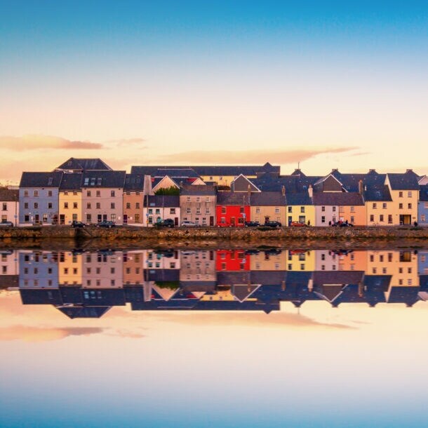 Bunte Häuser in Galway, Irland, entlang eines ruhigen Wassers mit Spiegelung. Die Gebäude sind in verschiedenen Farben, darunter Rot, Gelb und Weiß.