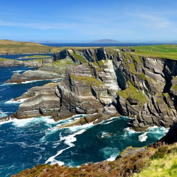 Luftaufnahme einer Küstenlandschaft mit steilen Klippen, grünem Gras und einem schmalen Weg, der entlang der Klippen verläuft. Im Hintergrund sind sanfte Hügel und ein blauer Himmel zu sehen.