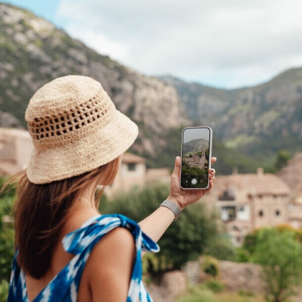 Frau mit Strohhut und blauem Kleid fotografiert Bergdorf mit Smartphone