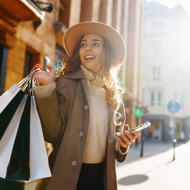 Eine junge Frau mit Hut steht in einer Innenstadt und hält ein Smartphone und mehrere Einkaufstüten in den Händen.