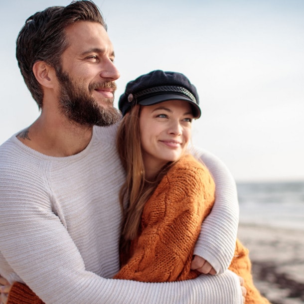 Ein sich umarmendes Paar in Pullovern steht glücklich lächelnd an einem Sandstrand und blickt aufs Meer.