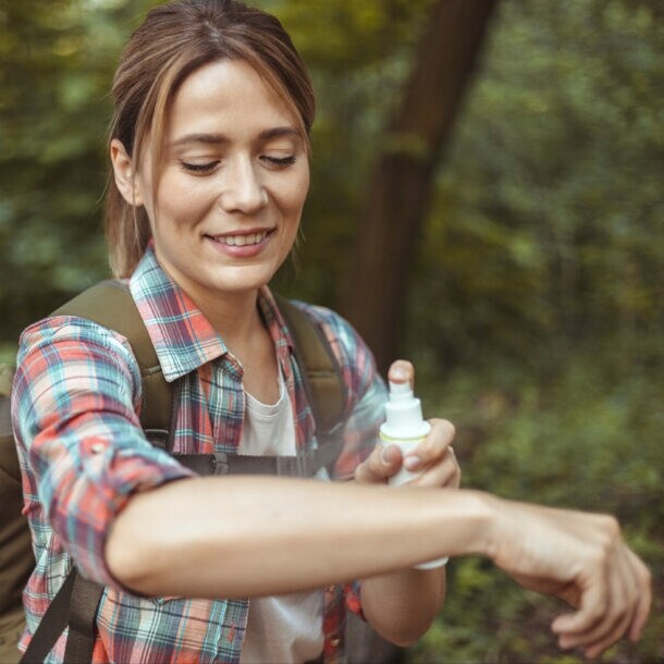 Person mit kariertem Hemd und Rucksack trägt im Wald Insektenschutzspray auf den Arm auf.