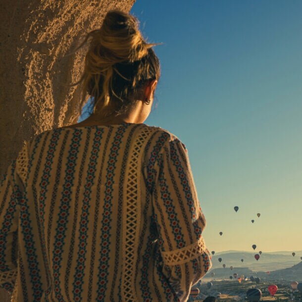 Eine Person mit einem langen, gemusterten Oberteil steht mit dem Rücken zur Kamera und blickt auf eine Landschaft mit vielen Heißluftballons im Himmel bei Sonnenaufgang.