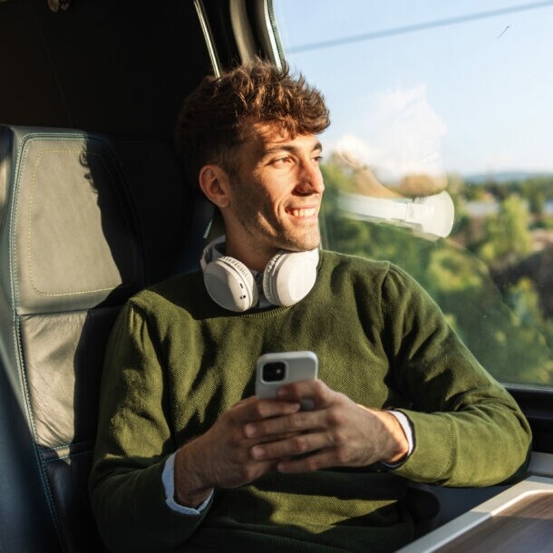 Junger Mann mit grünem Pullover und weißen Kopfhörern sitzt im Zug mit Smartphone in der Hand und blickt aus dem Fenster.