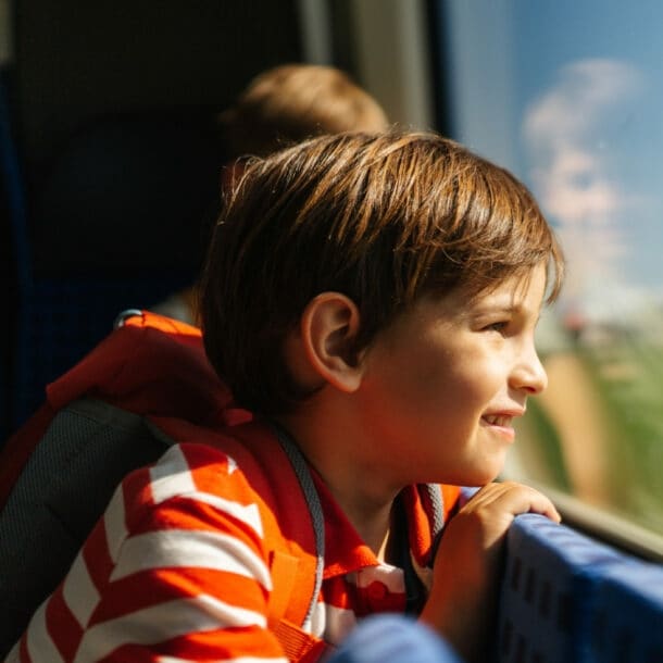 Ein Kind mit gestreiftem Oberteil lehnt sich am Zugfenster und schaut nach draußen. Im Hintergrund ist ein weiteres Kind zu sehen.