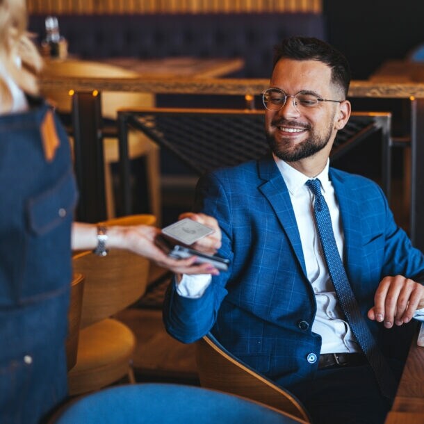 Ein Mann im blauen Anzug sitzt an einem Tisch in einem Bistro und hält eine Kreditkarte an ein Kartenlesegerät, das eine Person ihm hinhält.
