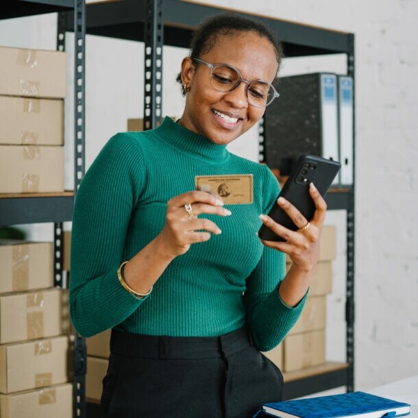 Eine lächelnde Frau im grünen Shirt steht vor einem Lagerregal mit Kartons und hält ein Smartphone sowie eine goldene Kreditkarte von American Express vor sich in den Händen.