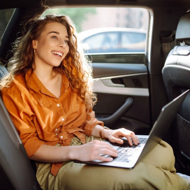Eine lachende Frau in orangefarbener Bluse sitzt auf dem Rücksitz eines Autos mit einem aufgeklappten Laptop auf ihrem Schoß.